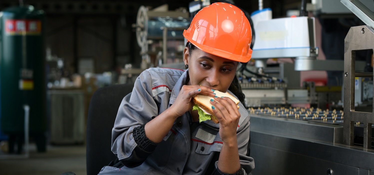 A young black woman in an orange helmet eats a sandwich in a production facility, next to a conveyor belt. Production worker taking a lunch break at the workplace