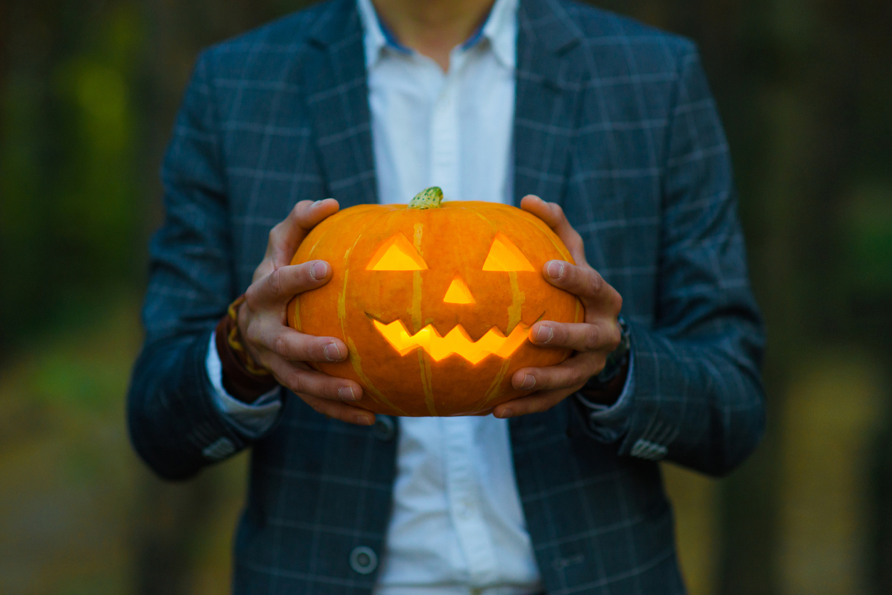 Man in suit holds a carved glowing pumpkin lantern with a creepy face. Halloween concept